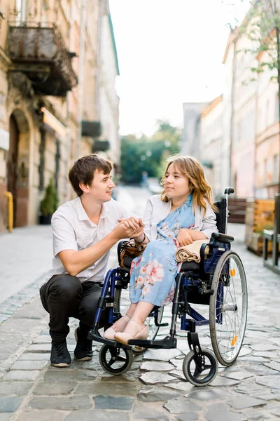 Handsome man and pretty woman in wheelchair looking at each other on street. Lovely couple in wheechair walking together in the city — 스톡 사진