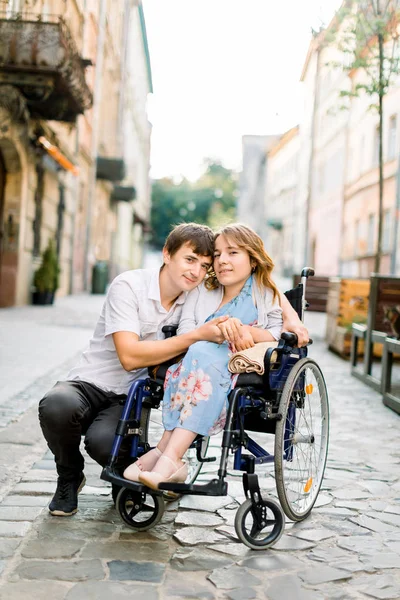 Hübsche junge lächelnde Frau im Rollstuhl und gutaussehender Mann, der in die Kamera schaut, Händchen haltend, verliebt, in der Altstadt spazierend. — Stockfoto