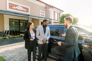 Otomobil işi, araba satışı, teknoloji ve insan konsepti - iş çifti, Afrikalı erkek ve beyaz kadın araba satıcısı açık hava otomobil salonunun bahçesinde siyah arabanın yanında duruyor.