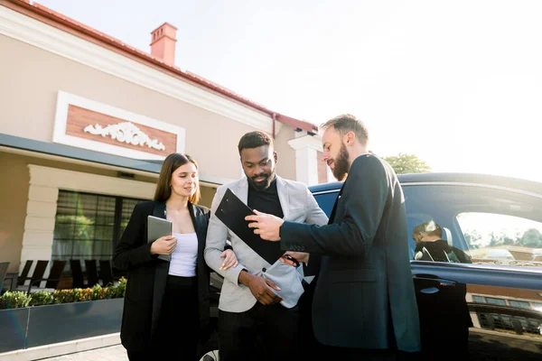 İş çifti, Afrikalı erkek ve beyaz kadın dışarıda siyah arabanın yanında genç adam satış müdürüyle duruyorlar. Aile arabayı satın alıyor. Sözleşmeyi gösteren zarif asistan.