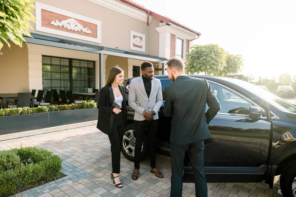 successful multiethnical business people standing outdoors on the territory of car dealership. Young man manager showing the car to the couple, African guy and Caucasian girl. Sale or rent of cars