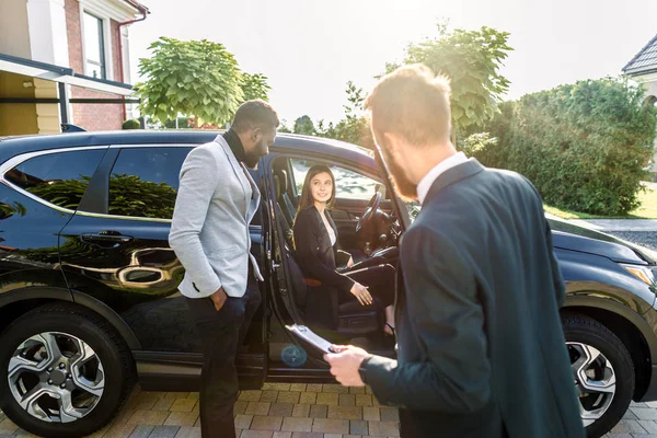 Young business couple, African man and Caucasian woman, looking for car to buy, woman sitting n the car, man standing and smiling. Back view of sales dealer holding the contract — 스톡 사진