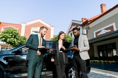 Üç başarılı iş adamı, Afrikalı ve Kafkasyalı erkek ve beyaz kadın, dışarıda arabanın yanında dururken tablet ve dizüstü bilgisayar kullanıyorlar..
