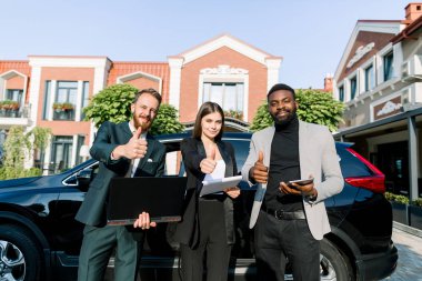 Çok ırklı başarılı bir ekip, iki işadamı ve bir iş kadını, elinde tablet, laptop ve kağıtlar var, baş parmaklarını kaldırıyor, dışarıda arabanın yanında duruyorlar. Takım, iş, teknoloji kavramı