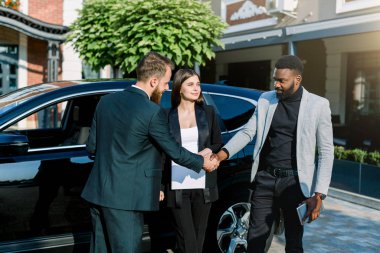 İki yakışıklı adam, Afrikalı ve Kafkasyalı, arabanın önünde dururken gülümseyerek el sıkışıyorlar. Güzel beyaz kadın aralarında duruyor..
