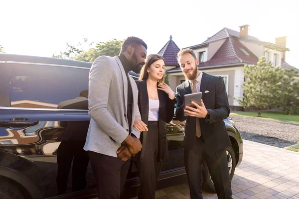 Agent d'assurance ou de vente avec contrat de remplissage de comprimés ou formulaire d'assurance près de voiture neuve noire et parler avec les clients, couple d'affaires, homme africain et femme caucasienne — Photo