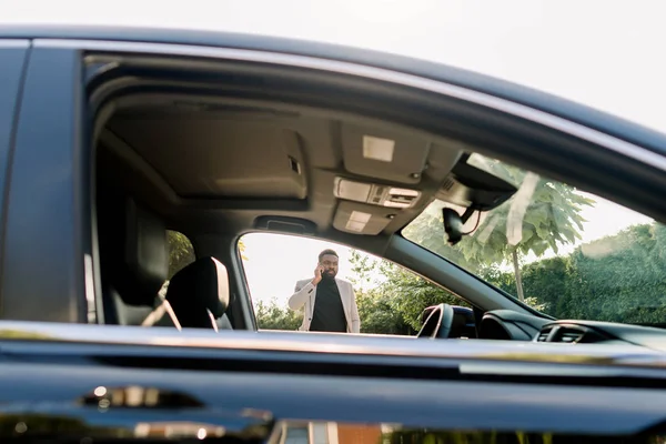 Sidovy av den svarta bilen fönstret och afrikanska affärsman i kostym talar med smartphone på bakgrunden — Stockfoto