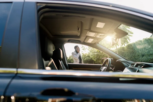 Photo of the car window on sunny day and successful African businessman walking while taking at the phone on the background. African american businessman in a suit speaking on smartphone. — 스톡 사진