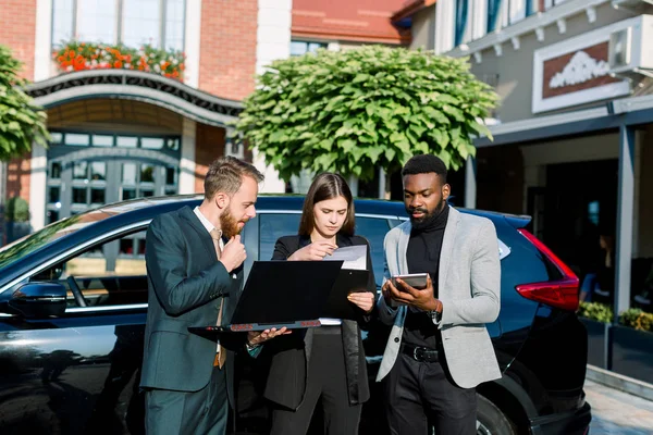 Jeunes trois collègues multiraciaux d'affaires utilisant ordinateur portable et tablette debout à l'extérieur dans le centre d'affaires près de la nouvelle voiture noire . — Photo