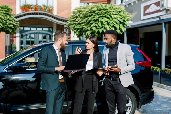 Groupe d'affaires multiethnique, deux hommes et une femme qui utilisent ou regardent la tablette et l'ordinateur portable et discutent du travail et du projet debout près de la voiture noire crossover prête pour la réunion. Concept d'entreprise . — Photo