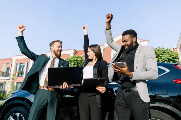 Ομάδα χαρούμενων πολυεθνικών επιχειρηματιών που εργάζονται με φορητό υπολογιστή και tablet και γιορτάζουν την επιτυχία, δείχνοντας ενθουσιασμό υψώνοντας γροθιές, ενώ στέκεται έξω μπροστά από το αυτοκίνητο. έννοια του επιχειρηματικού ομίλου — Φωτογραφία Αρχείου