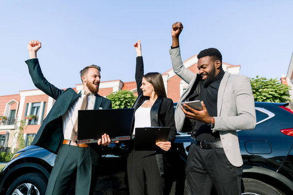 Group of happy multiethnic business people working with laptop and tablet and celebrate success, showing excite by raise fists up, while standing outdoors in front of the car. business group concept