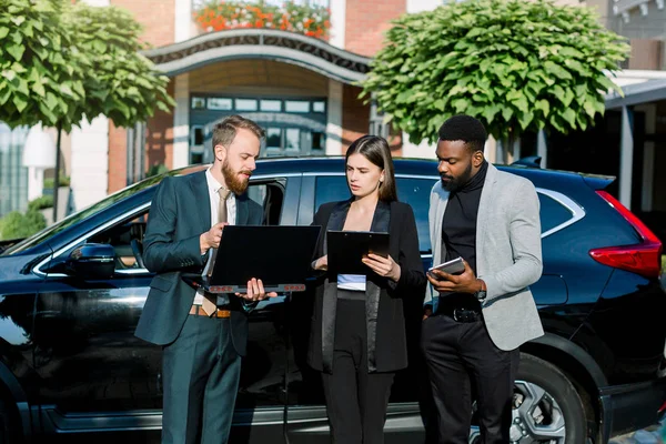 Les gens d'affaires, l'homme africain, l'homme et la femme caucasiens, travaillant avec ordinateur portable et tablette, tout en se tenant à l'extérieur du bureau près de la voiture — Photo