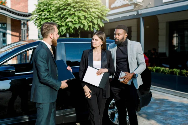 Les gens d'affaires se réunissent dehors. Image de jeunes partenaires d'affaires multiraciaux attrayants debout près de la voiture noire — Photo