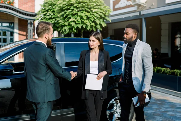 Les gens d'affaires se serrent la main, finissant une réunion. Homme et femme caucasiens serrant la main, homme africain collègue se tient debout et tient la tablette. Rencontre à l'extérieur, près de la voiture — Photo