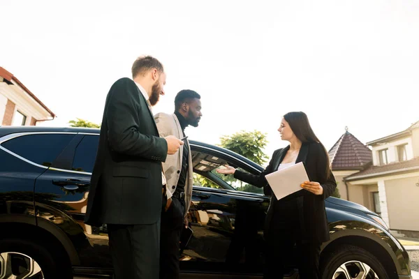 Jeunes gens d'affaires, deux hommes, caucasiens et africains, debout à la cour du salon de voiture en plein air avec une jeune vendeuse ou un gestionnaire achetant ou louant une nouvelle voiture noire crossover — Photo