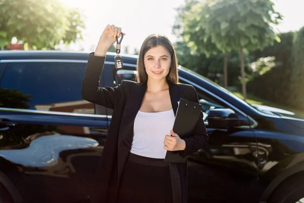 Beautiful girl with car key in hand. Caucasian woman car seller holding car keys, standing in front of new black car outdoors in vehicle trade fair. Auto rental or sales concept. — 스톡 사진