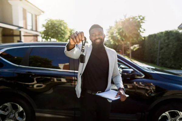 Servicio de alquiler de coches. Happy African Man gerente de ventas o cliente sosteniendo la llave y smilikng Cerca de nuevo coche negro. Enfoque en clave — Foto de Stock