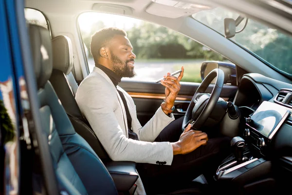 Pebisnis muda tampan Afrika duduk di mobil baru yang mahal memegang ponsel di tangannya. Mobil dan konsep bisnis — Stok Foto