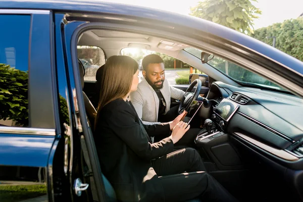 Young African American man communicating with female Caucasian coworker during car drive to business meeting discussing startup ideas and presentation from digital tablet — 스톡 사진