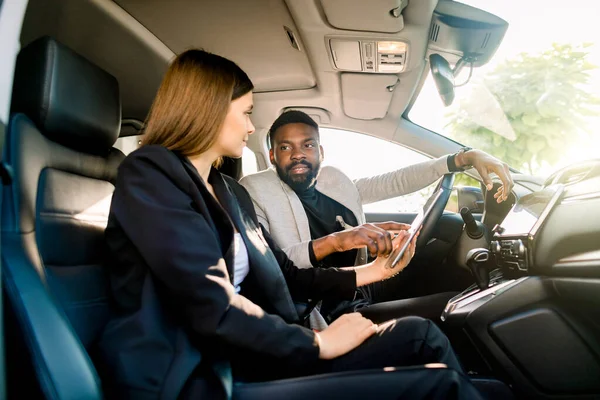 Pareja de negocios, bonita mujer caucásica y apuesto hombre africano, trabajando en coche con tableta digital. El hombre muestra algo en la tableta para mujer. Negocios, finanzas, concepto de venta de coches — Foto de Stock