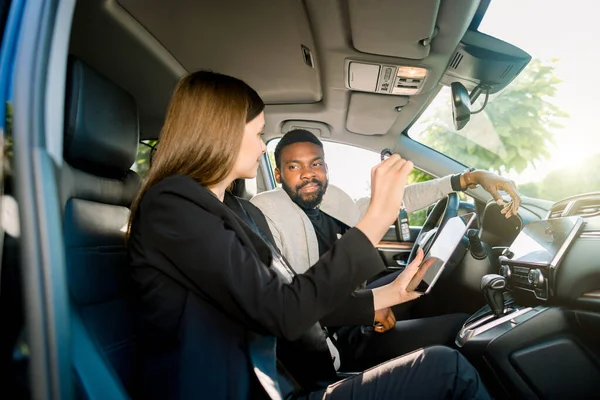 Car sale and rent, people concept. Happy African man and Caucasian woman car dealer with tablet computer sitting in new car. Woman salesmanager holds car keys and showing the rent contract on tablet — 스톡 사진