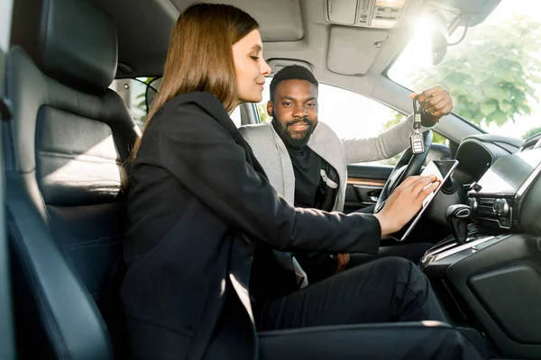 Snygg glad ung afrikansk man som håller bilnycklar från sin nya lyxbil, medan du sitter i bilen tillsammans med söt kvinna bilförsäljaren håller en tablett — Stockfoto