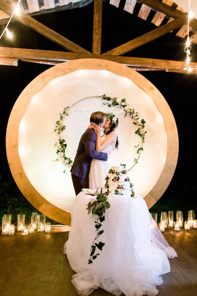 Bride and a groom kissing, standing behind their rustic wedding cake on banquet. Rustic wedding reception outdoors in wooden tent — Stock Photo, Image
