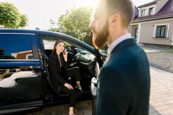 Ung vacker affärskvinna kommer ut genom ytterdörren i sin bil. Förare eller affärspartner hjälper kvinnan att komma ut ur bilen — Stockfoto