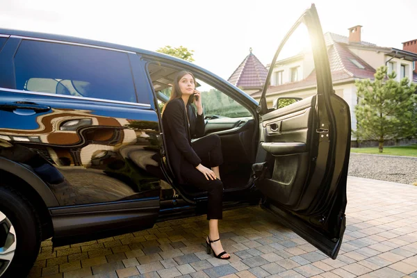 Beautiful business woman in dark suit is using a smart phone and smiling while sitting on passenger seat in the luxury black car — 스톡 사진