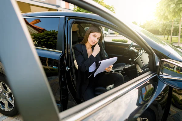 Ung affärskvinna talar i telefon i passagerarsätet i bilen och hålla i handen en Urklipp med papper för att skriva anteckningar — Stockfoto