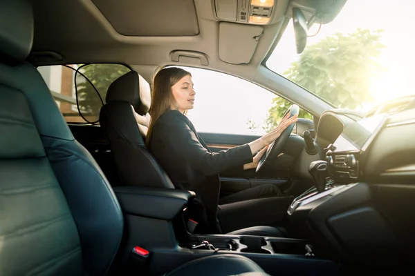 Beautiful young brunette woman in business suit driving a car to office. Businesswoman drive a car in city. Side view, sunny day — 스톡 사진