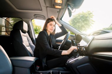 Şoför koltuğunda oturan ve kameraya bakan genç ve güzel iş kadınının yan görüntüsü. Güneşli bir gün. Arabadaki iş kadını