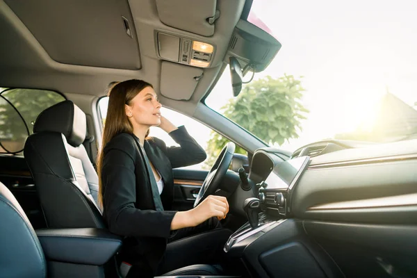 Vacker ung affärskvinna i svart kostym tittar i backspegeln på en bil. Ung leende affärskvinna ser sig själv i backspegeln. Sidovy — Stockfoto