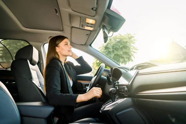 Georgeous young business woman in black suit looking in the rearview mirror of a car. Young smiling businesswoman looking herself in rearview mirror. Side view — 스톡 사진