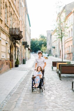 Tekerlekli sandalyedeki güzel genç kadın yakışıklı erkeğiyle şehir merkezinde yürüyüş yaparken eğleniyor.