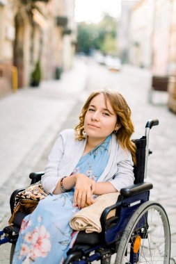 Mavi elbiseli, arka planda tekerlekli sandalyede oturan, gülümseyen, engelli bir kadının yakın plan portresi.