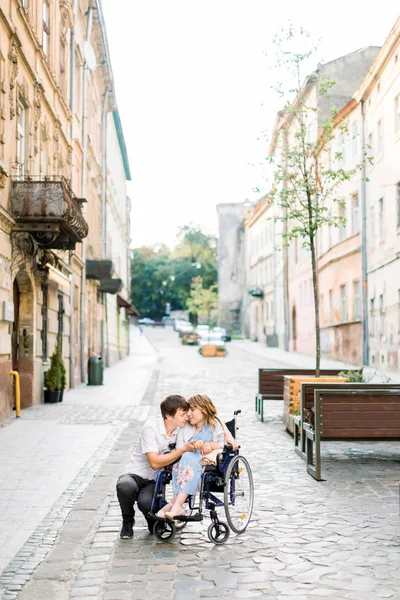 Закохана пара в старому центрі міста. Красива молода дівчина з хворобою на інвалідному візку і її мила людина, що йде, посміхається і торкається лоба, закриті очі — стокове фото