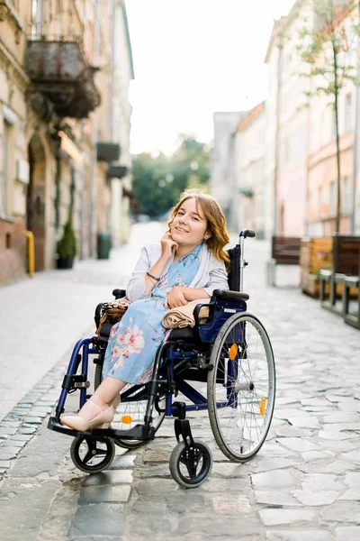 Ελκυστική κοπέλα με ειδικές ανάγκες ξανθιά κοπέλα με μπλε φόρεμα κάθεται σε μια αναπηρική καρέκλα, σε εξωτερικούς χώρους στην πόλη — Φωτογραφία Αρχείου