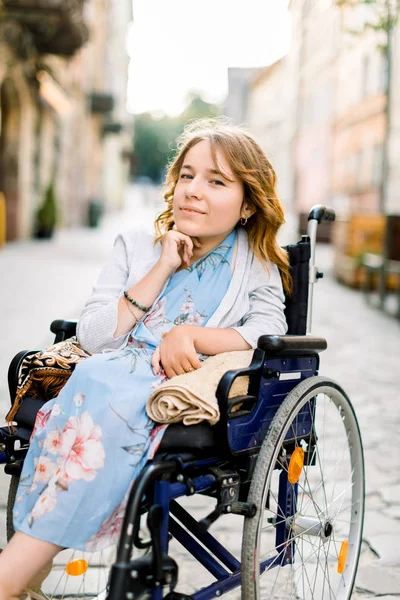 Κοντινό πλάνο πορτρέτο της όμορφης νεαρής χαμογελαστής γυναίκας με μπλε φόρεμα, που κάθεται σε αναπηρική καρέκλα στο φόντο των κτιρίων της παλιάς πόλης — Φωτογραφία Αρχείου
