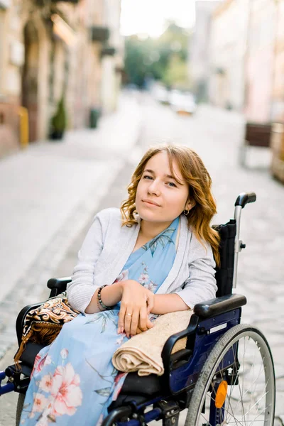 Κοντινό πλάνο πορτρέτο της όμορφης νεαρής χαμογελαστής γυναίκας με μπλε φόρεμα, που κάθεται σε αναπηρική καρέκλα στο φόντο των κτιρίων της παλιάς πόλης — Φωτογραφία Αρχείου