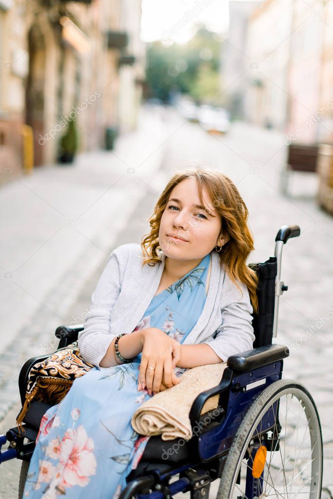 Closeup portrait of pretty young smiling disabled woman in blue dress, sitting in wheelchair on the background of old city buildings