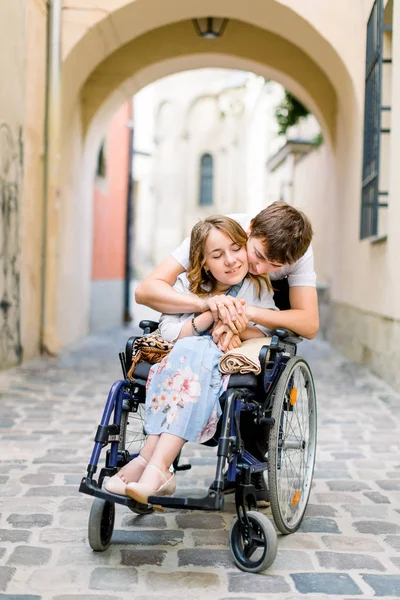 Verliebtes Paar in der Altstadt. hübsches junges Mädchen mit Krankheit im Rollstuhl und ihr liebenswerter Mann, der sie von hinten umarmt — Stockfoto