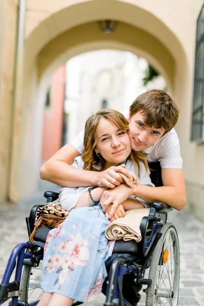 Ritratto ravvicinato di giovane donna felice in sedia a rotelle e suo marito, guardando la macchina fotografica e sorridendo, camminando all'aperto nella città vecchia — Foto Stock