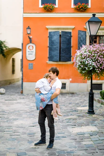 Mooie jonge gelukkig gehandicapte vrouw met haar knappe man, houdt haar vast aan de handen, terwijl ze buiten staat voor oude oranje gebouwen in de stad — Stockfoto