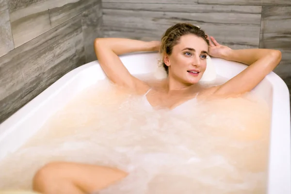 Mujer bastante joven está tumbada en el baño mirando de lado las manos detrás de la cabeza mientras recibe terapia de hidromasaje en el spa —  Fotos de Stock