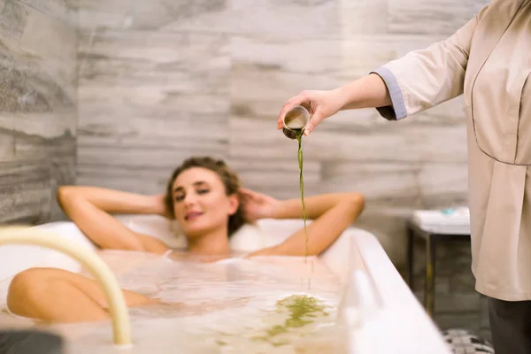 Widok z przodu młodej kobiety w kąpieli podczas hydromasażu w salonie piękności. Terapeutka wypełnia wannę specjalnym składnikiem do hydromasażu. Koncepcja pielęgnacji ciała i spa — Zdjęcie stockowe
