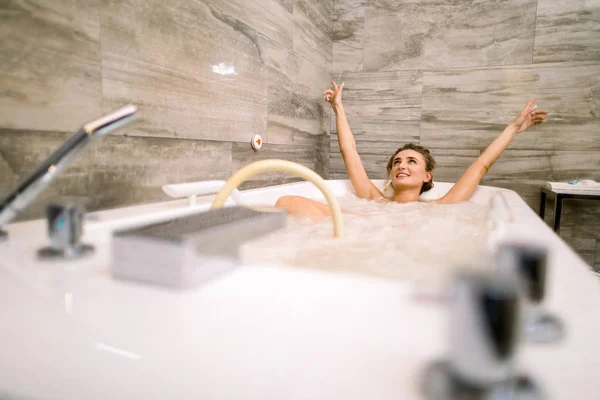 Piękna szczęśliwa uśmiechnięta kobieta ciesząca się i relaksująca w jacuzzi w centrum spa, mając profesjonalny hydromasaż i trzymając ręce do góry — Zdjęcie stockowe