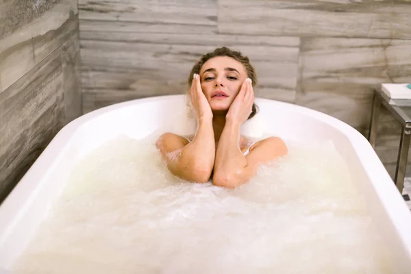 Retrato de cerca de una mujer joven relajándose en la bañera, recibiendo hidromasaje, tocando la cara —  Fotos de Stock