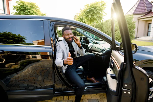 Beau et réussi homme d'affaires africain et patron assis dans la voiture, parlant au téléphone, souriant, regardant la caméra et montrant pouce vers le haut. Concept d'entreprise — Photo